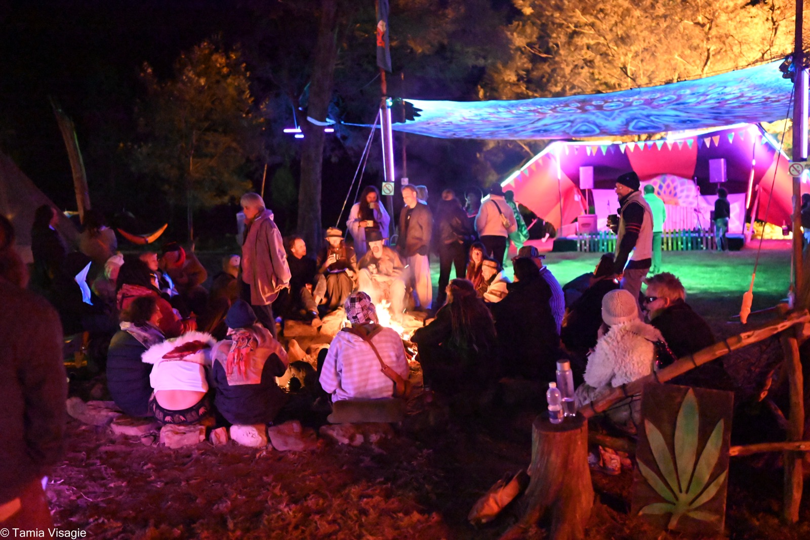 People gathered around a fire at night, a lit stage glowing behind them