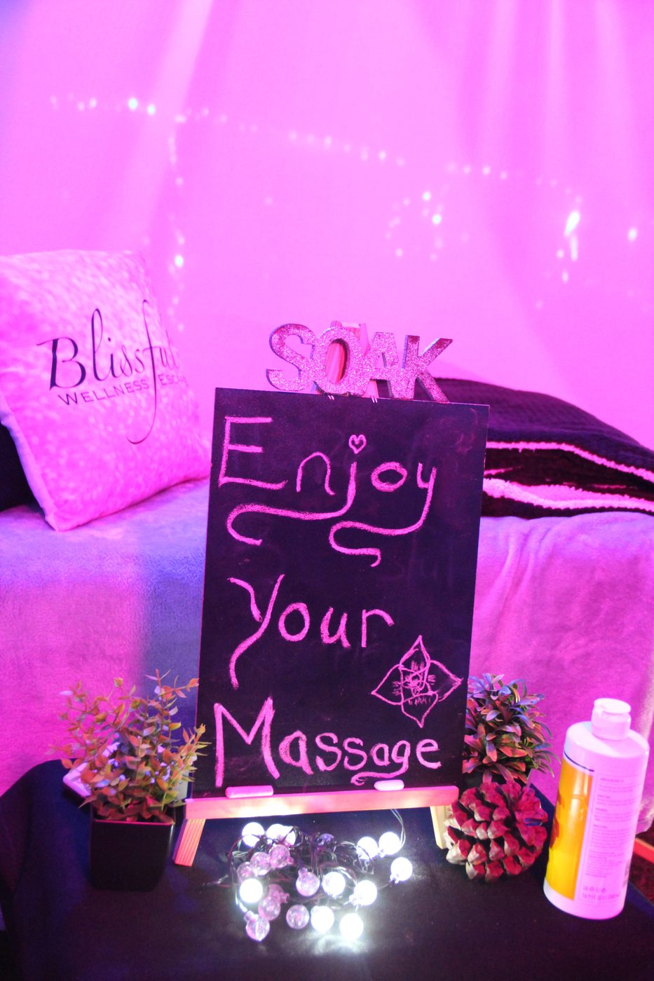 A pink-lit massage table with a chalkboard sign reading Enjoy Your Massage