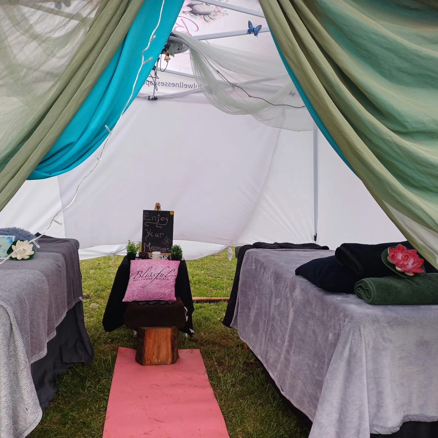 Two massage beds inside the draped tent with a chalkboard sign and a pink mat between them