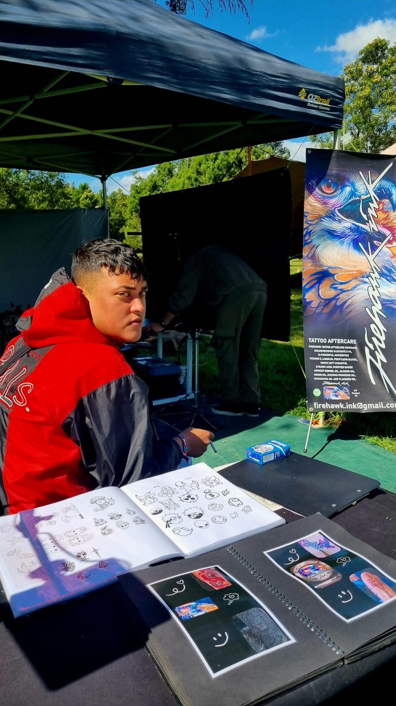 A Firehawk Ink artist in a red hoodie at the stall, with the flash book and sketch pages open on the table