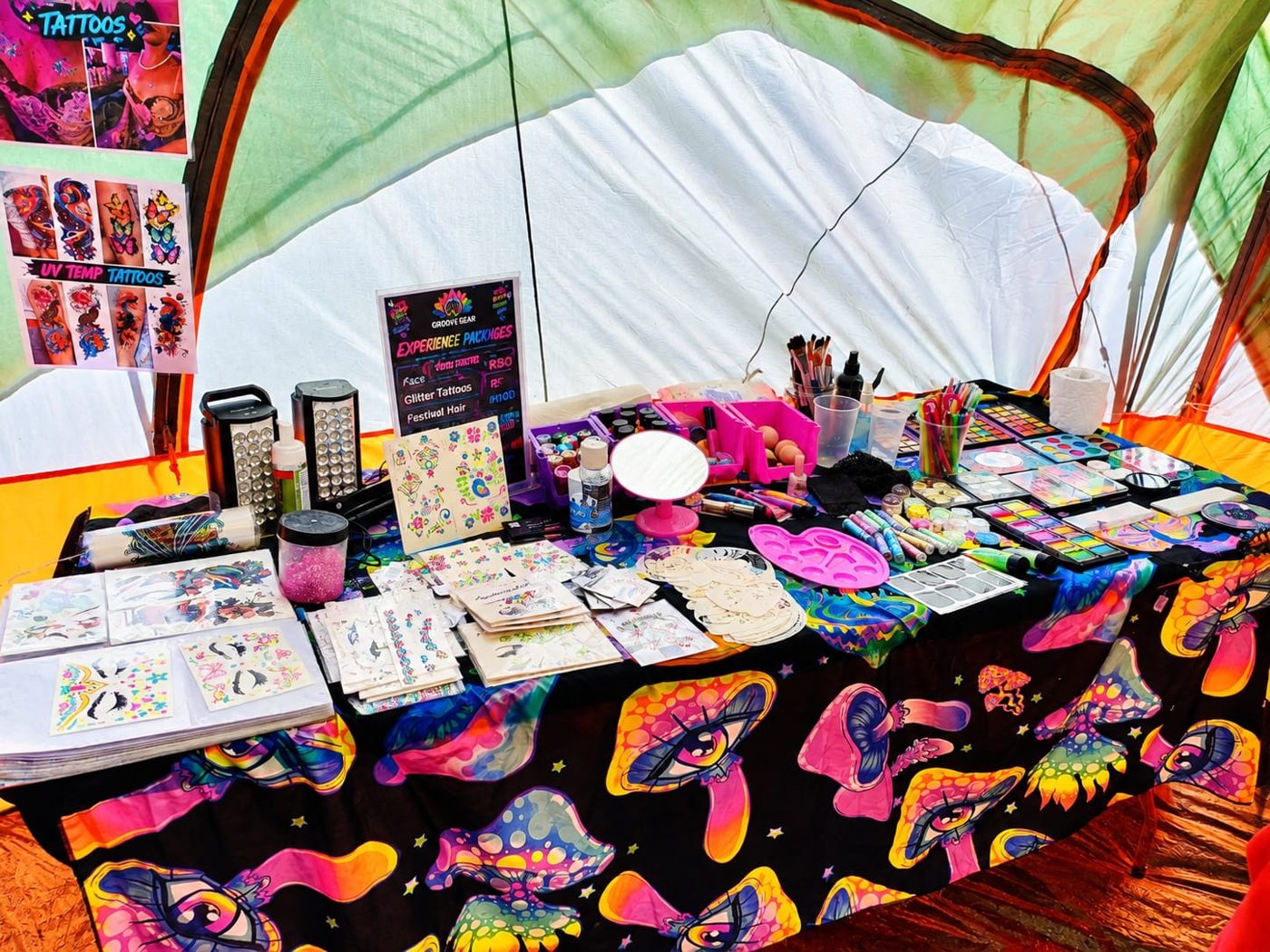 The body art station, with face paints, brushes and sheets of UV temporary tattoos on a mushroom-print tablecloth