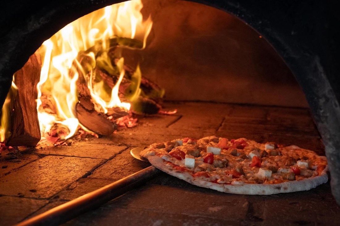 A pizza cooking on the wood-fired oven floor beside open flames