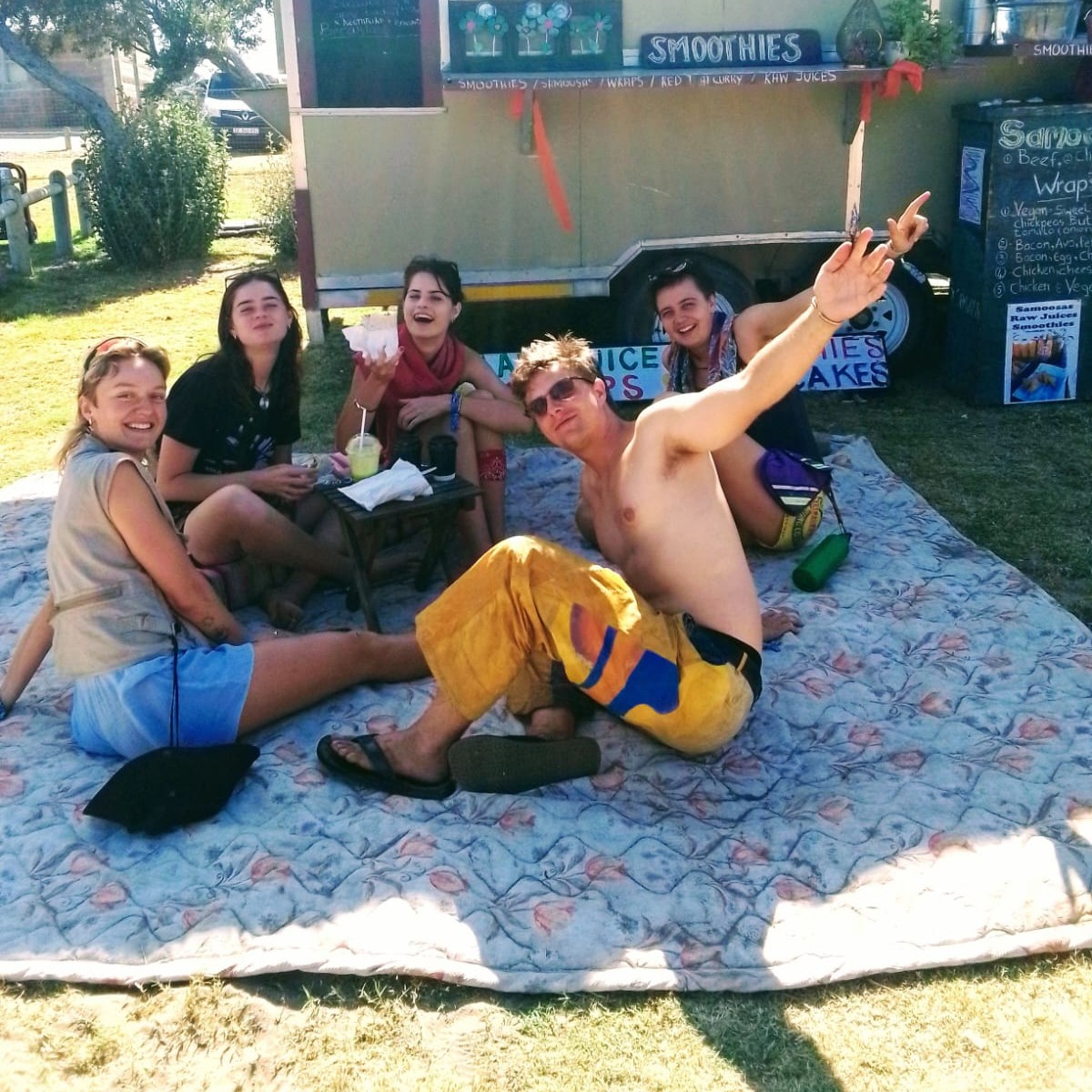 A group of friends laughing together on a picnic blanket in front of the Oh So Peachy trailer, smoothies in hand